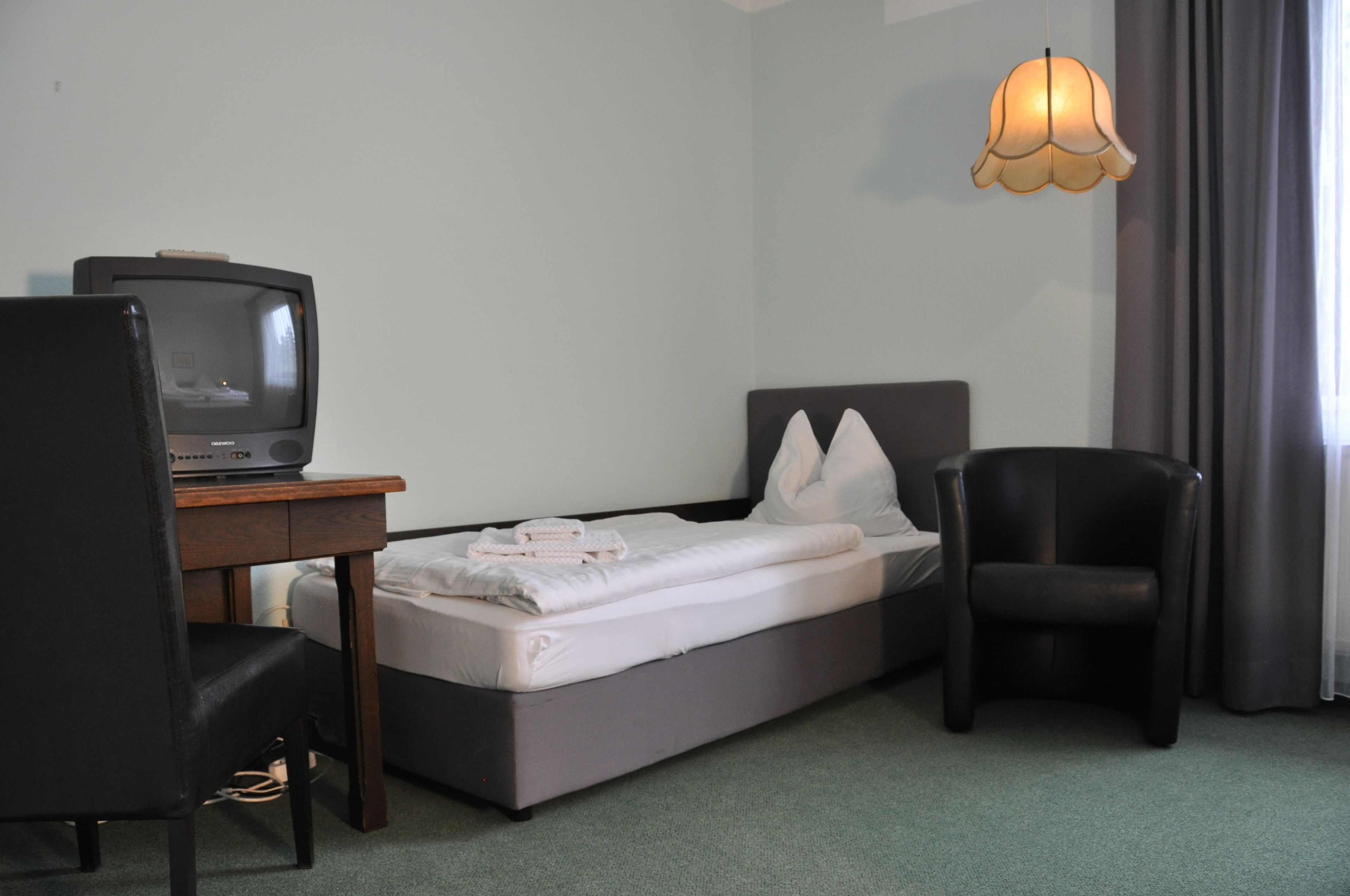 A hotel room with a single bed, a desk with a television, a chair, and a hanging light fixture.