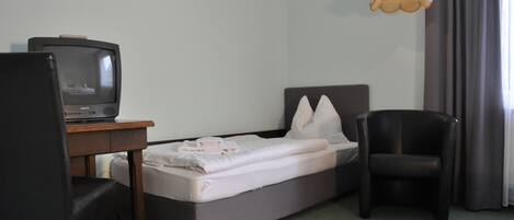 A hotel room with a single bed, a desk with a television, a chair, and a hanging light fixture.