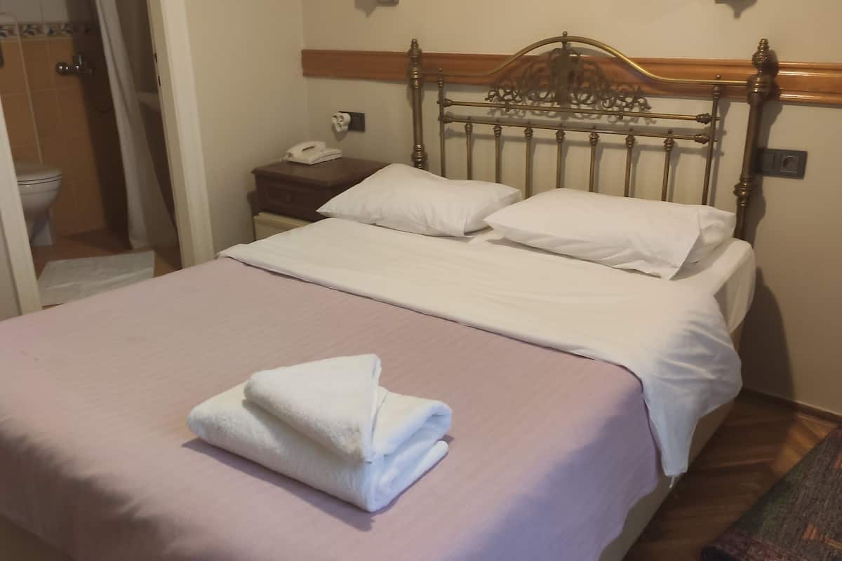 Ein ordentlich bezogenes Bett mit rosa Tagesdecke und weißen Leinentüchern, ein hölzernes Kopfteil, ein Nachttischchen mit Telefon und ein im Hintergrund sichtbares Badezimmer.