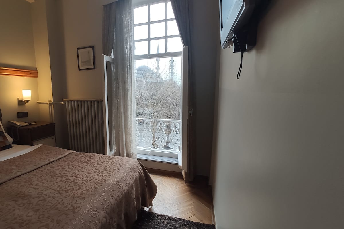 Ein Schlafzimmer mit Bett, Nachttisch, Lampe, Fenster mit Vorhängen, Heizkörper und einem an der Wand befestigten Fernseher.