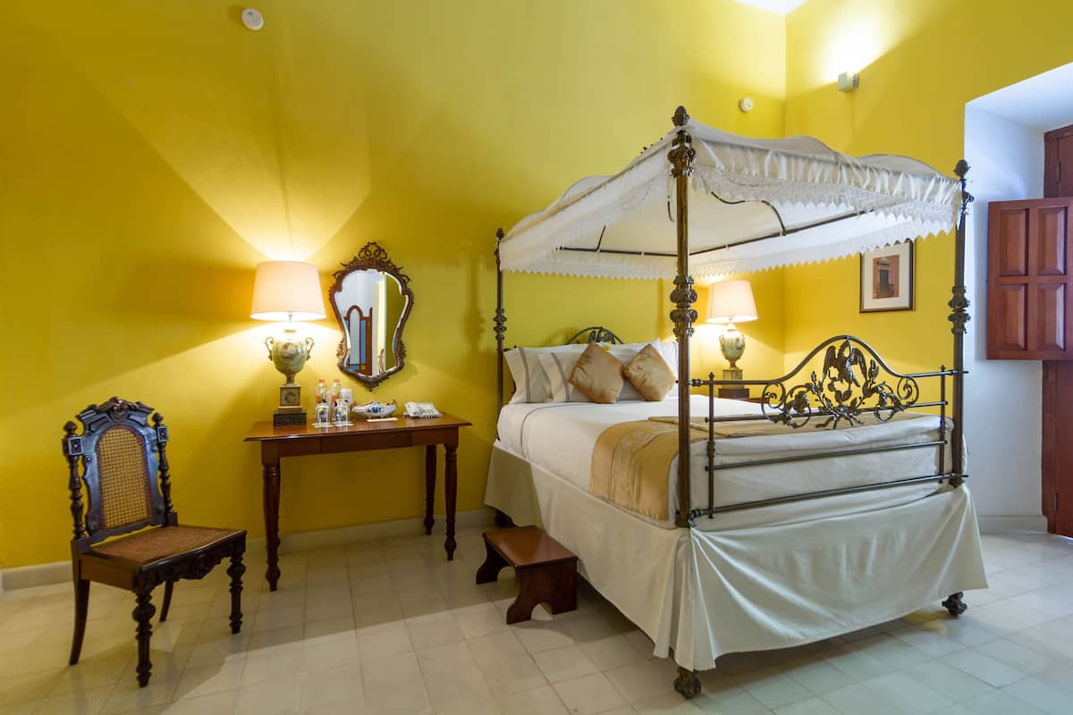 A four-poster bed with a canopy, a wooden nightstand with a lamp, a chair, and a small stool.
