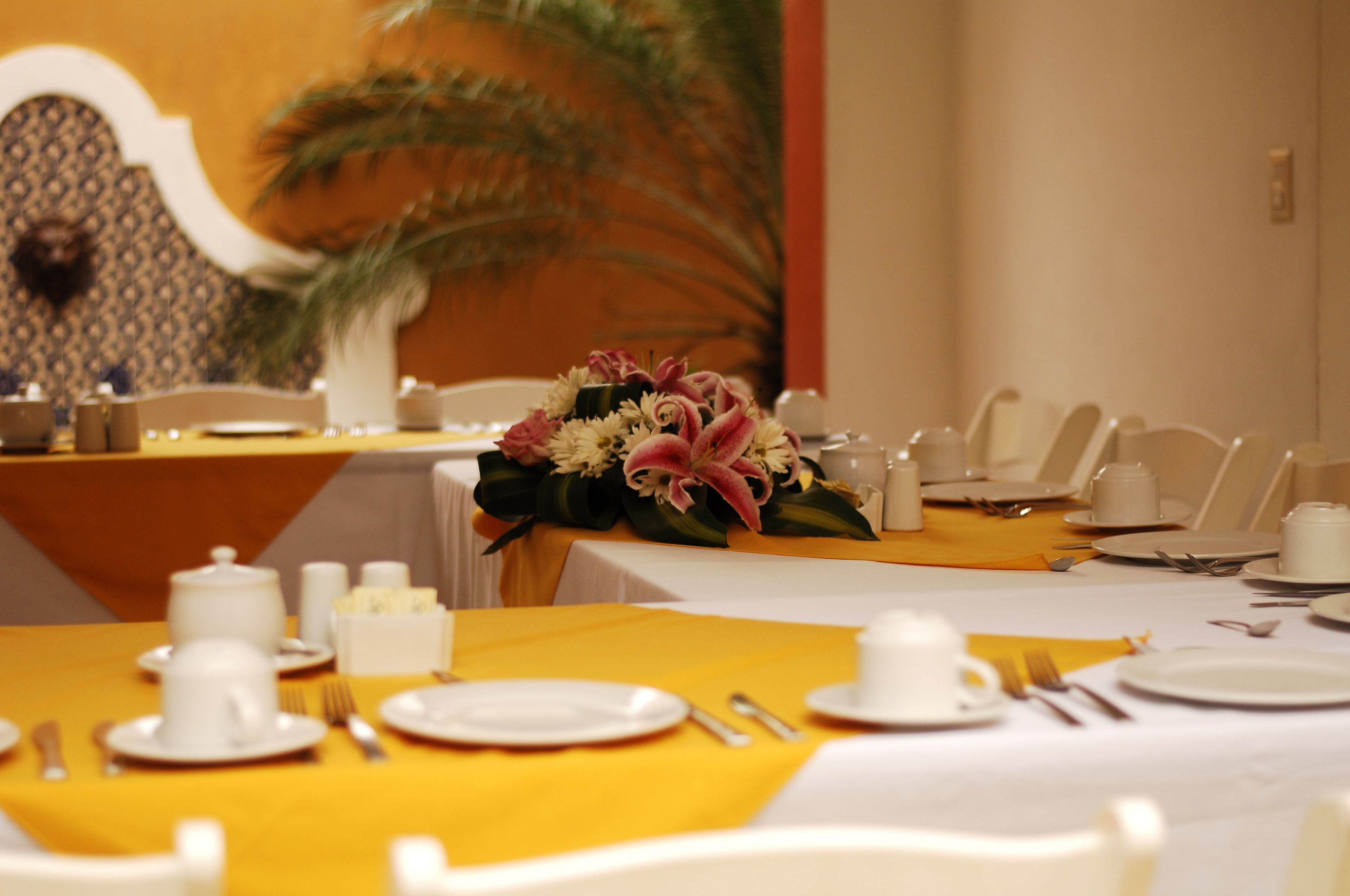 Un comedor con manteles amarillos, platos blancos y cubiertos. Hay un centro de mesa con flores sobre la mesa.