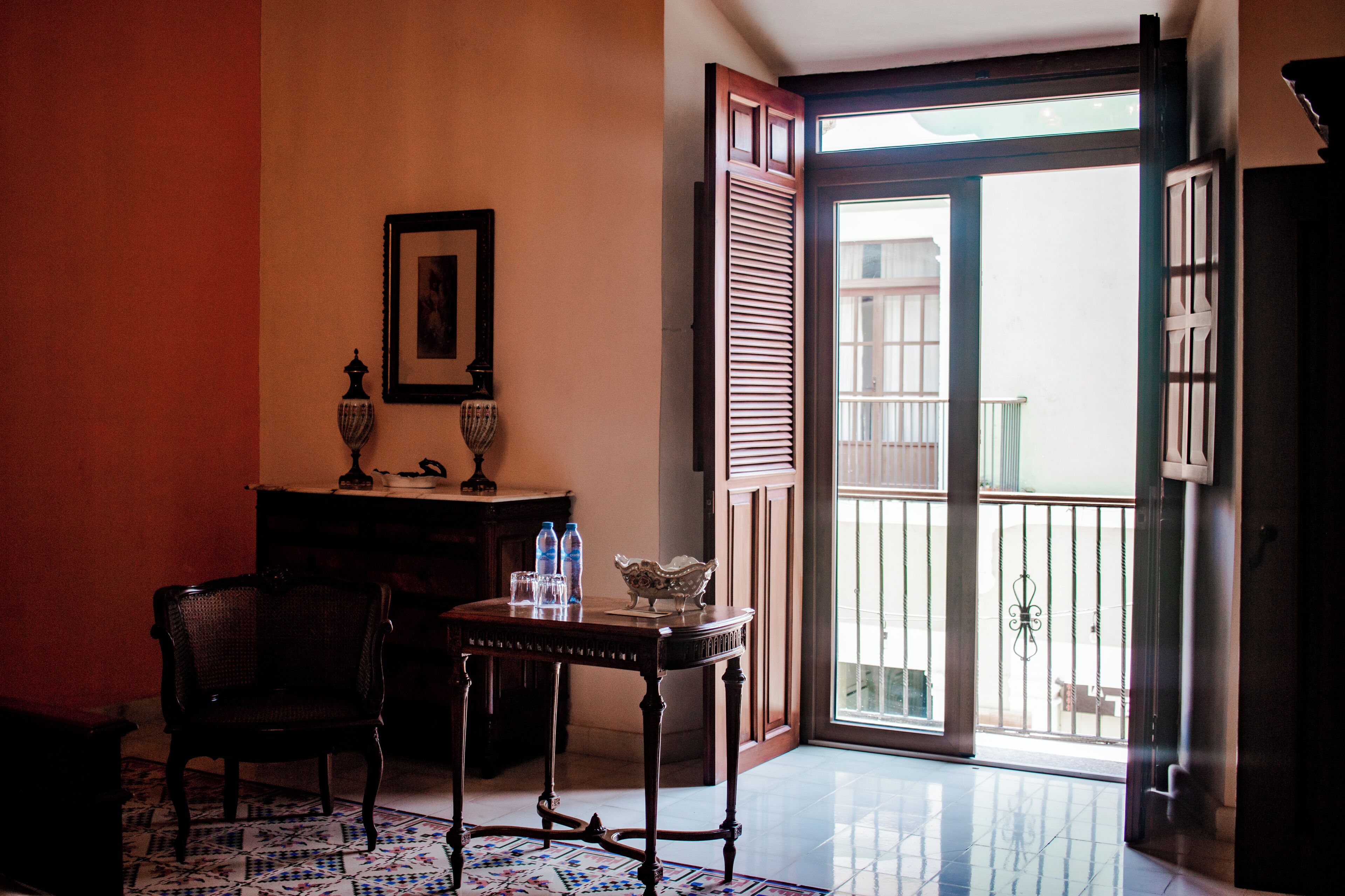 Una habitación con una mesa de madera, una silla, un tapete estampado, un cuadro enmarco y una puerta que da a un balcón.