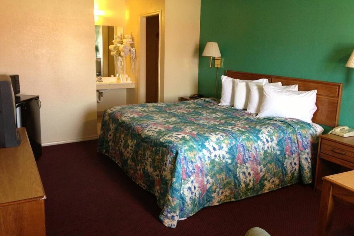 A hotel room with a floral bedspread, a wooden headboard, a desk with a lamp, and a mirror above the dresser.