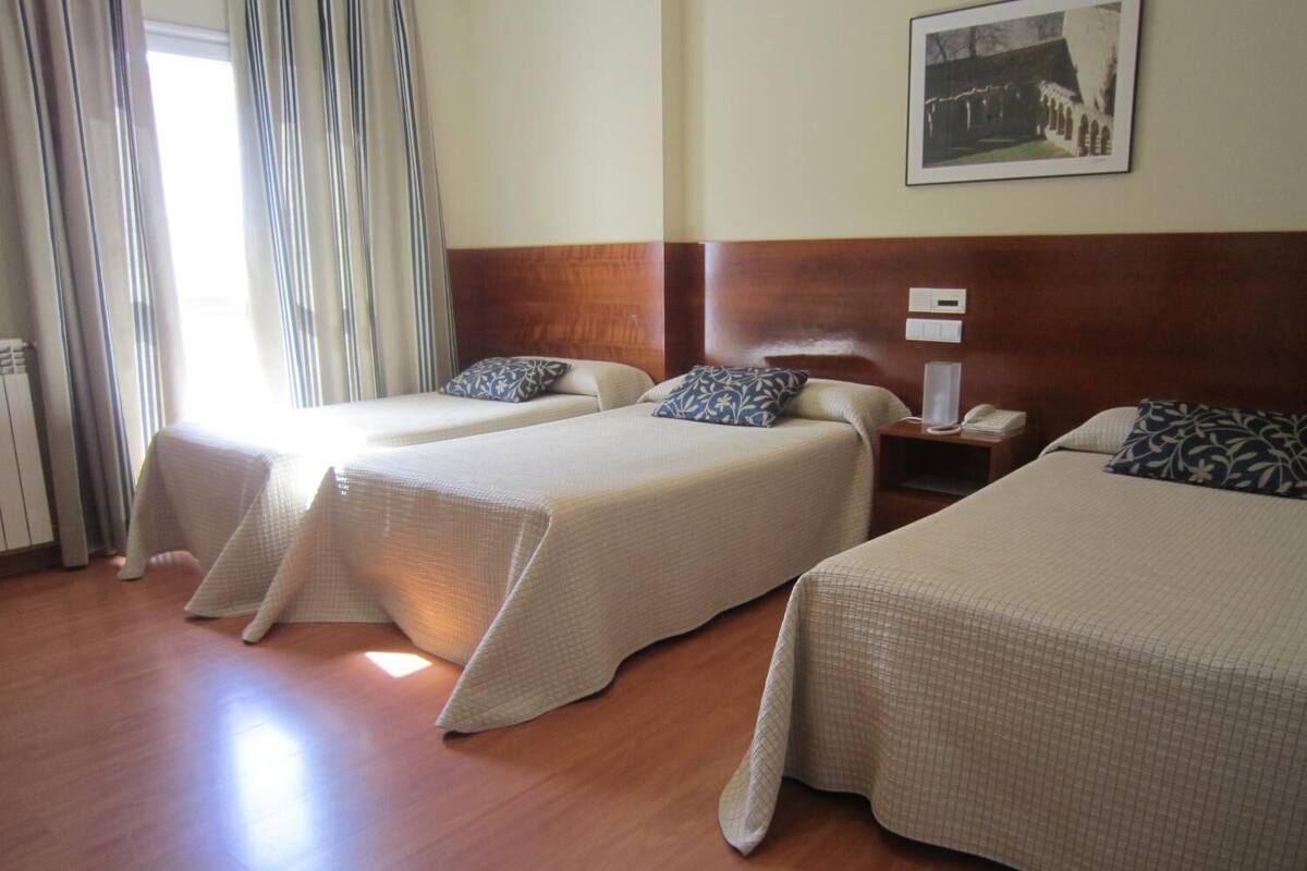 Ein Hotelzimmer mit drei Einzelbetten, einem hölzernen Kopfteil, einem Nachttisch mit Lampe und einem gerahmten Bild an der Wand.