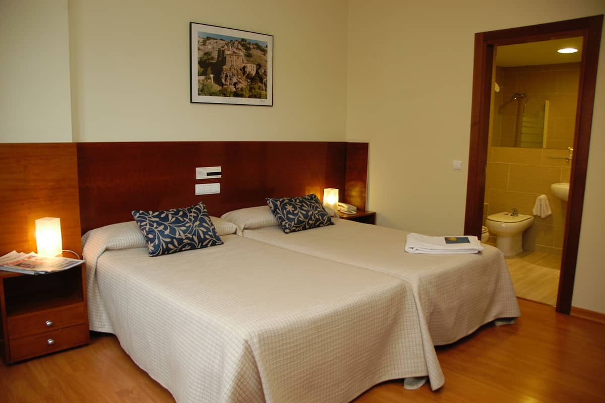 Ein Hotelzimmer mit zwei Betten, einem Nachttisch mit Lampe, einem gerahmten Bild an der Wand und einem durch eine offene Tür einsehbaren Badezimmer.