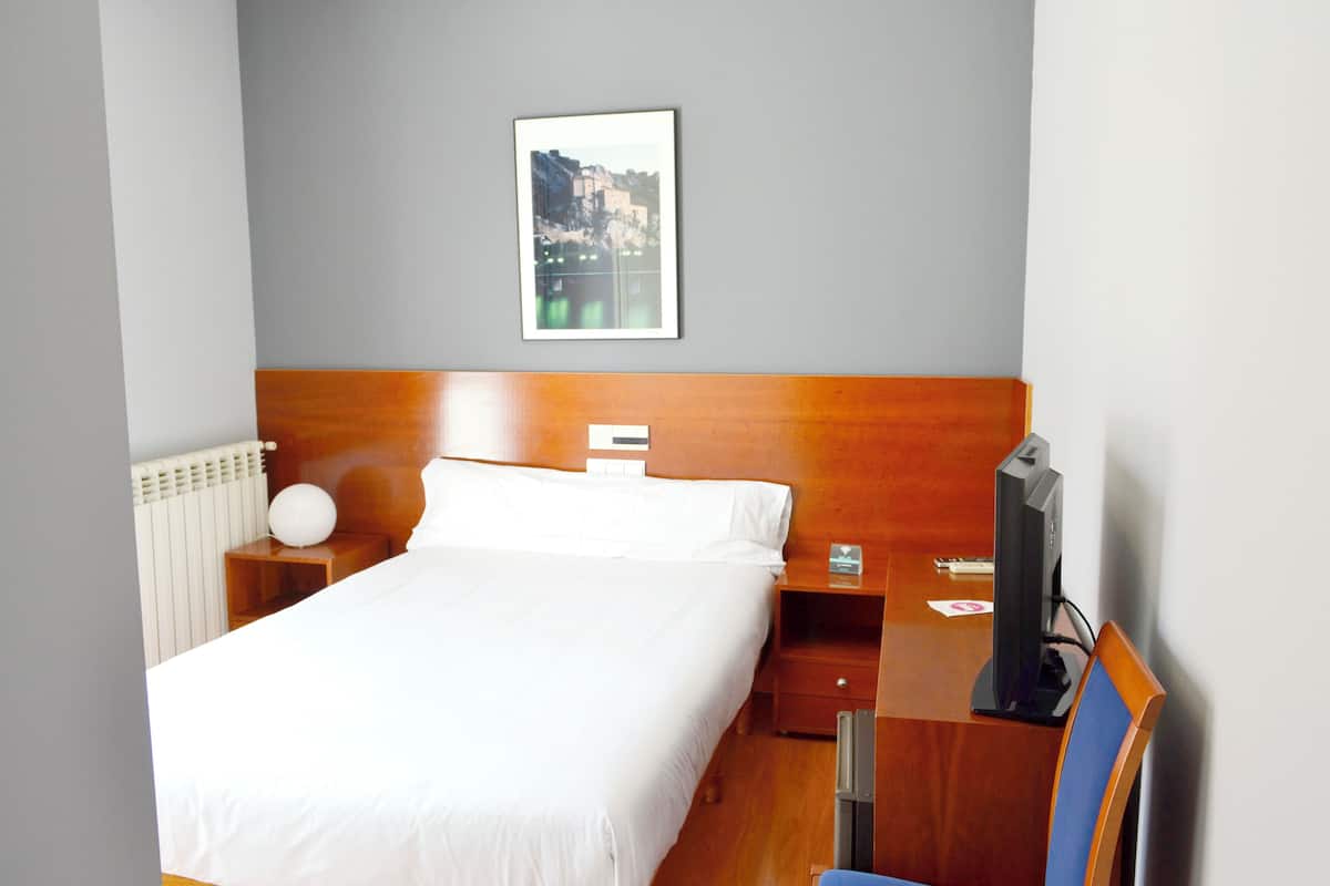 Ein Hotelzimmer mit Holz-Kopfteil, einem Bett mit weißer Bettwäsche, einem Nachttisch mit Lampe, einem Fernseher und einem gerahmten Bild an der Wand.