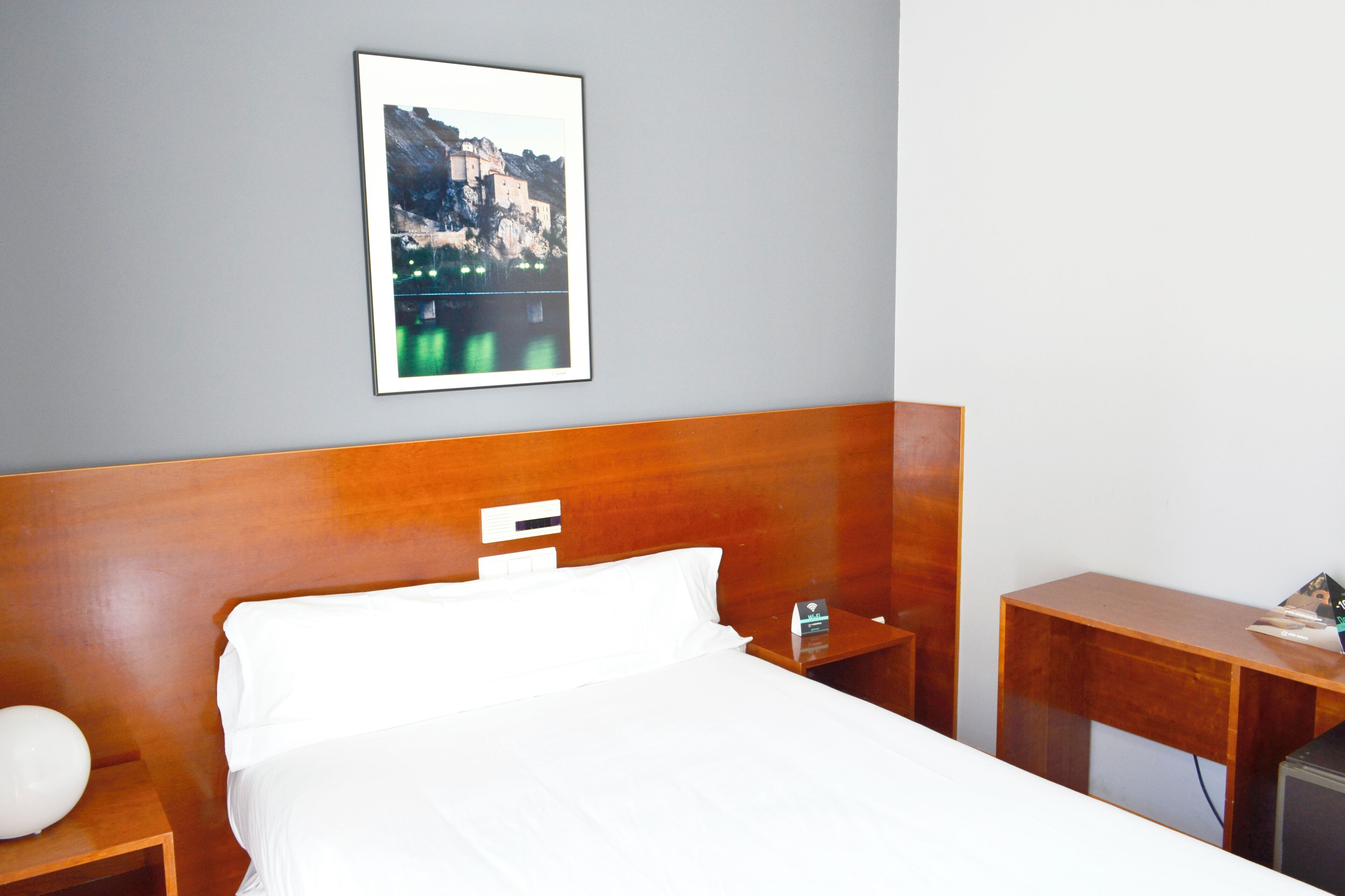 Una camera d'albergo con una testiera di legno, un letto con lenzuola bianche, un comodino con una lampada, un tavolino e un quadro appeso alla parete.