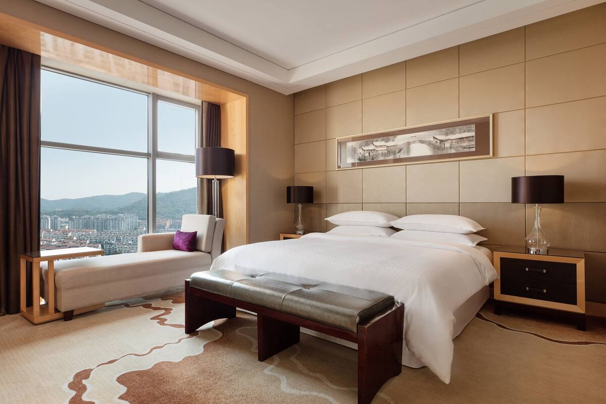 A modern hotel room with a large bed, a bench, a sofa, and a nightstand.