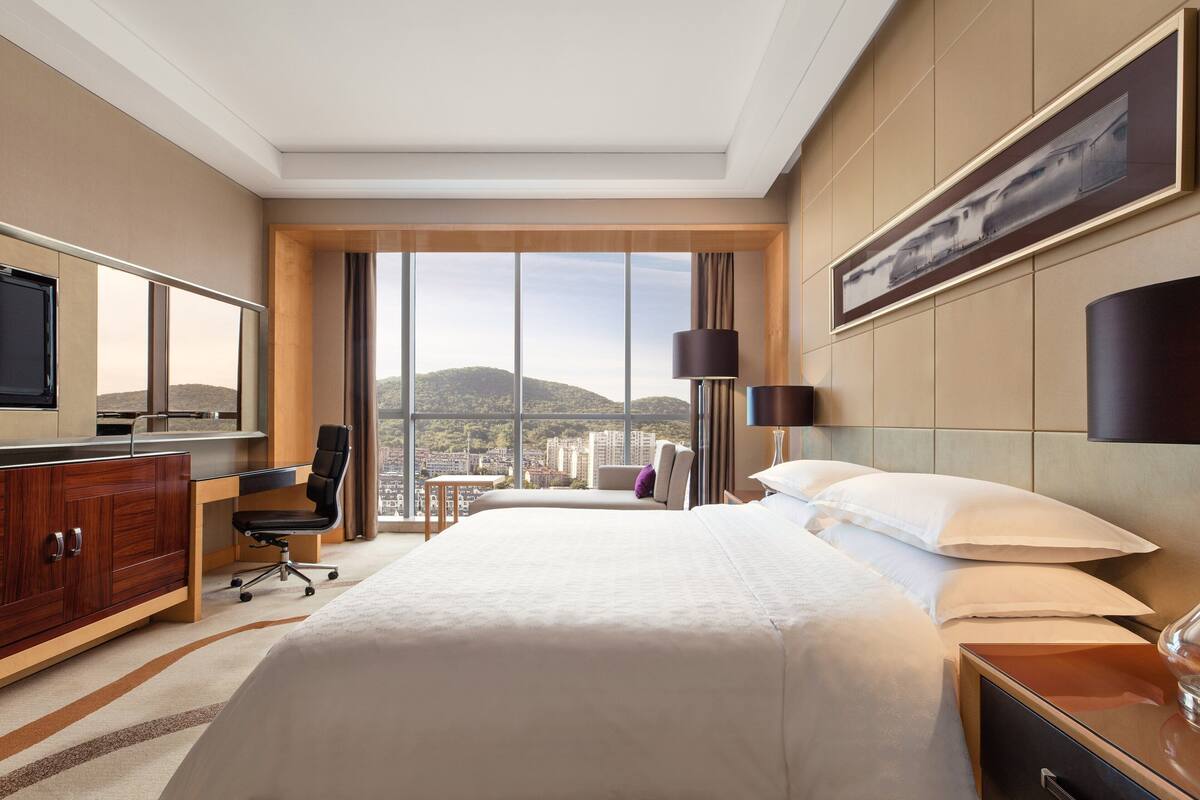 A hotel room with a large bed, a desk, a chair, a TV, and a view of the city.