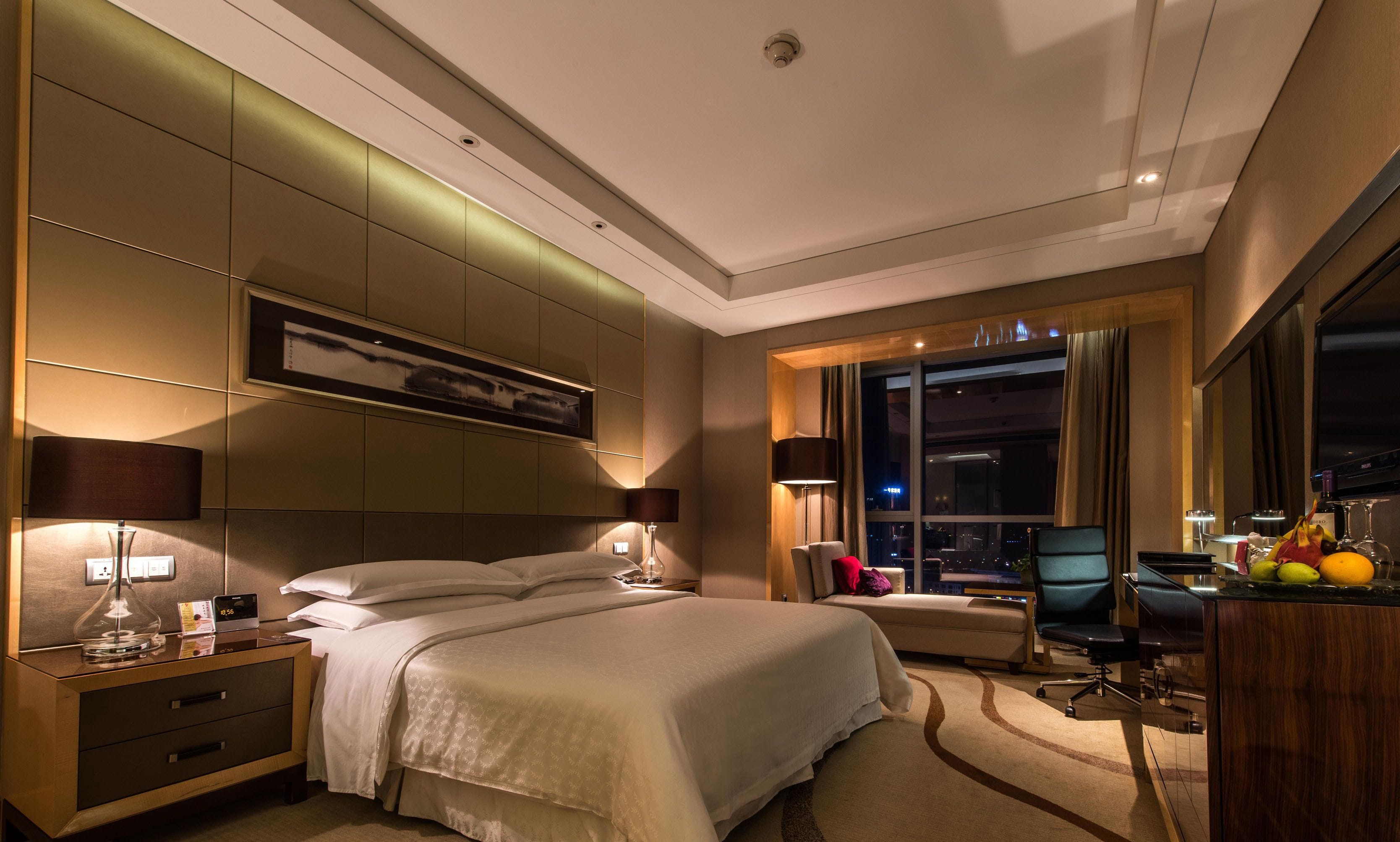 Una habitación de hotel moderna con una cama grande, mesitas de noche, un escritorio y vista a la ciudad de noche.