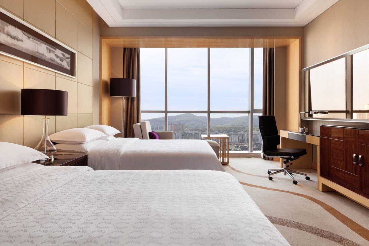 A hotel room with two beds, a desk, a chair, a lamp, and a large window offering a city view.