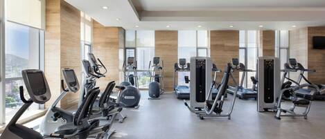 A modern gym with various cardio and strength training equipment, large windows, and a view of the city.