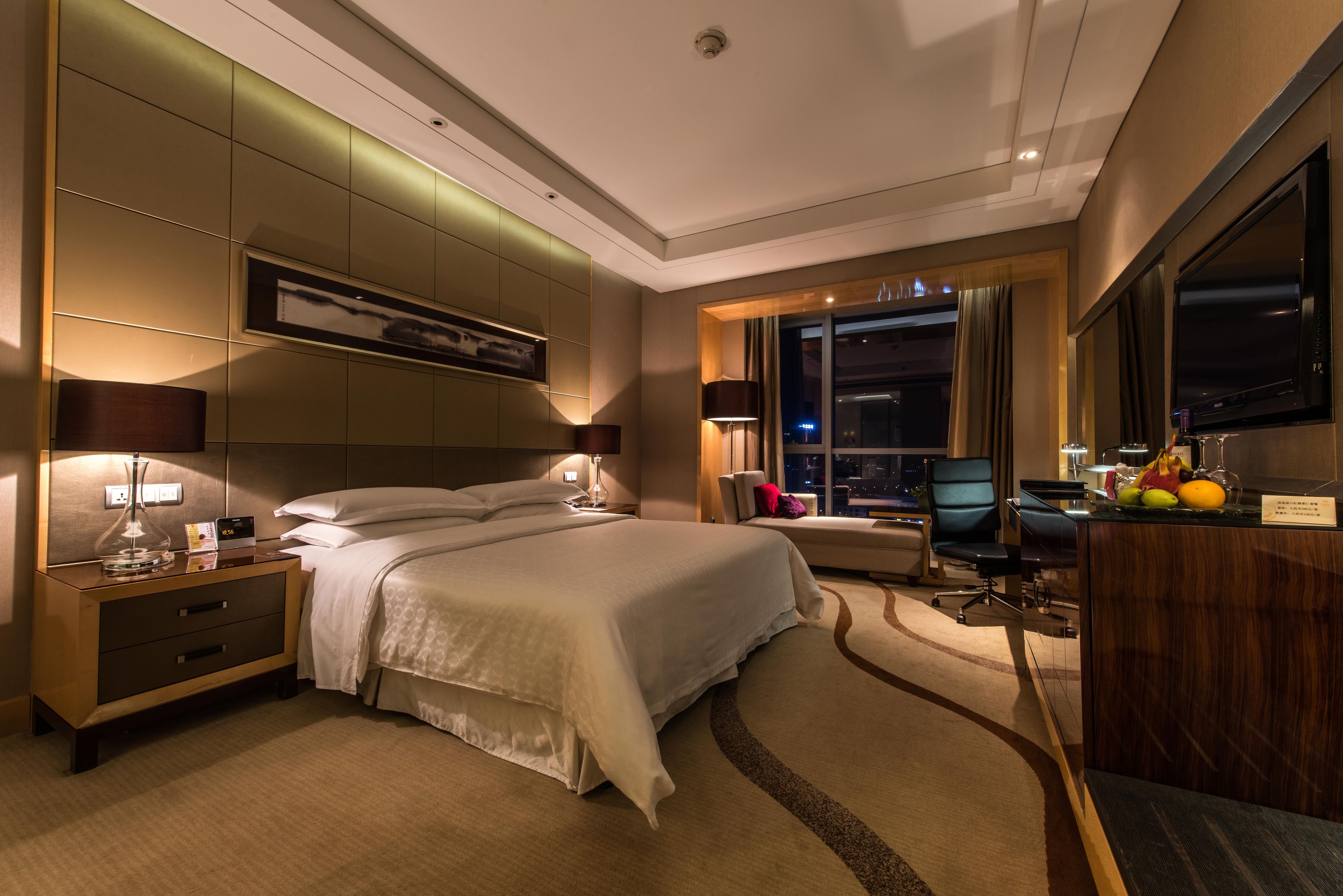 A modern hotel room with a large bed, a desk with a computer, and a view of the city at night.