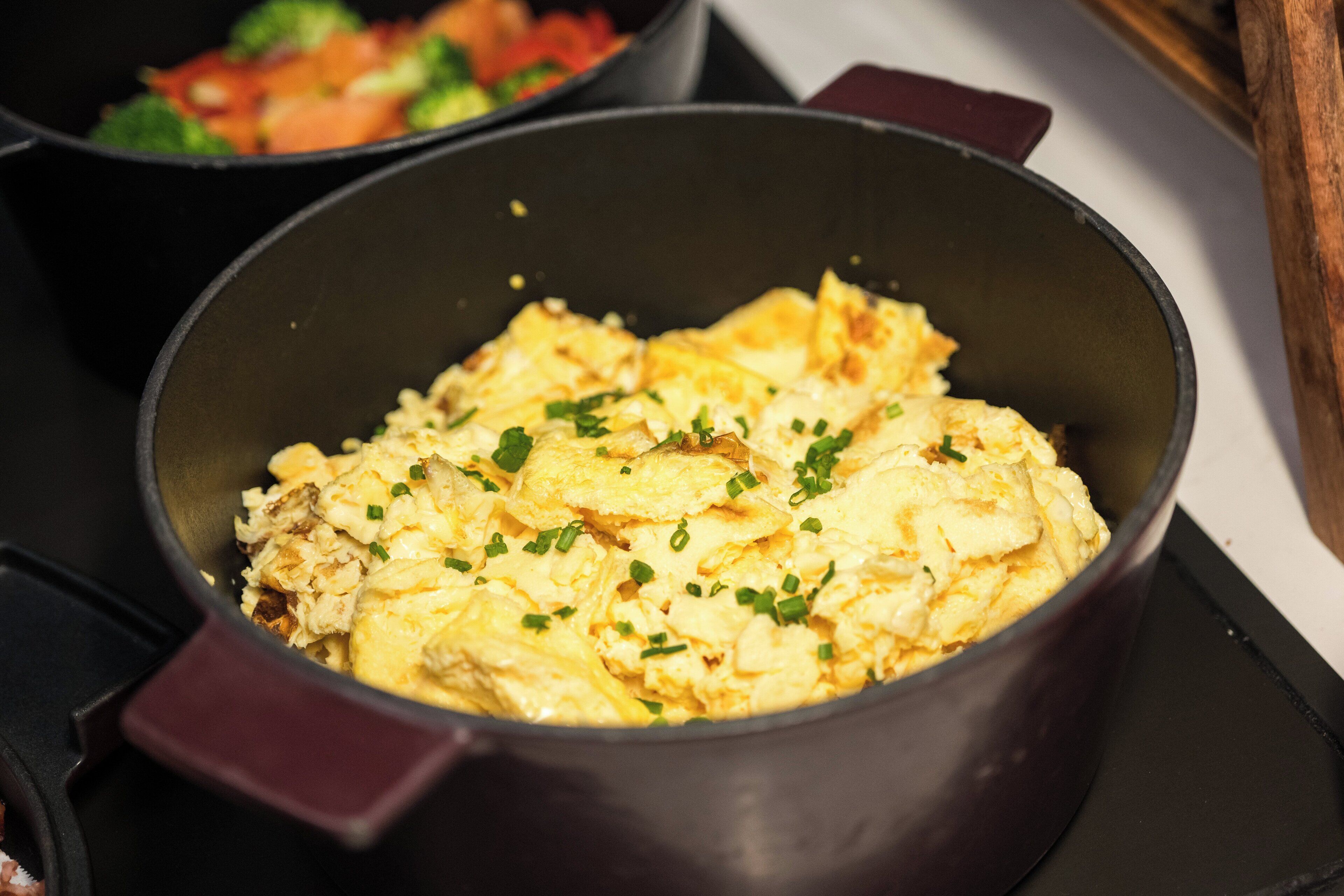 Primer plano de una olla con huevos revueltos adornados con cebollín, sobre un fondo desenfocado de una cocina.