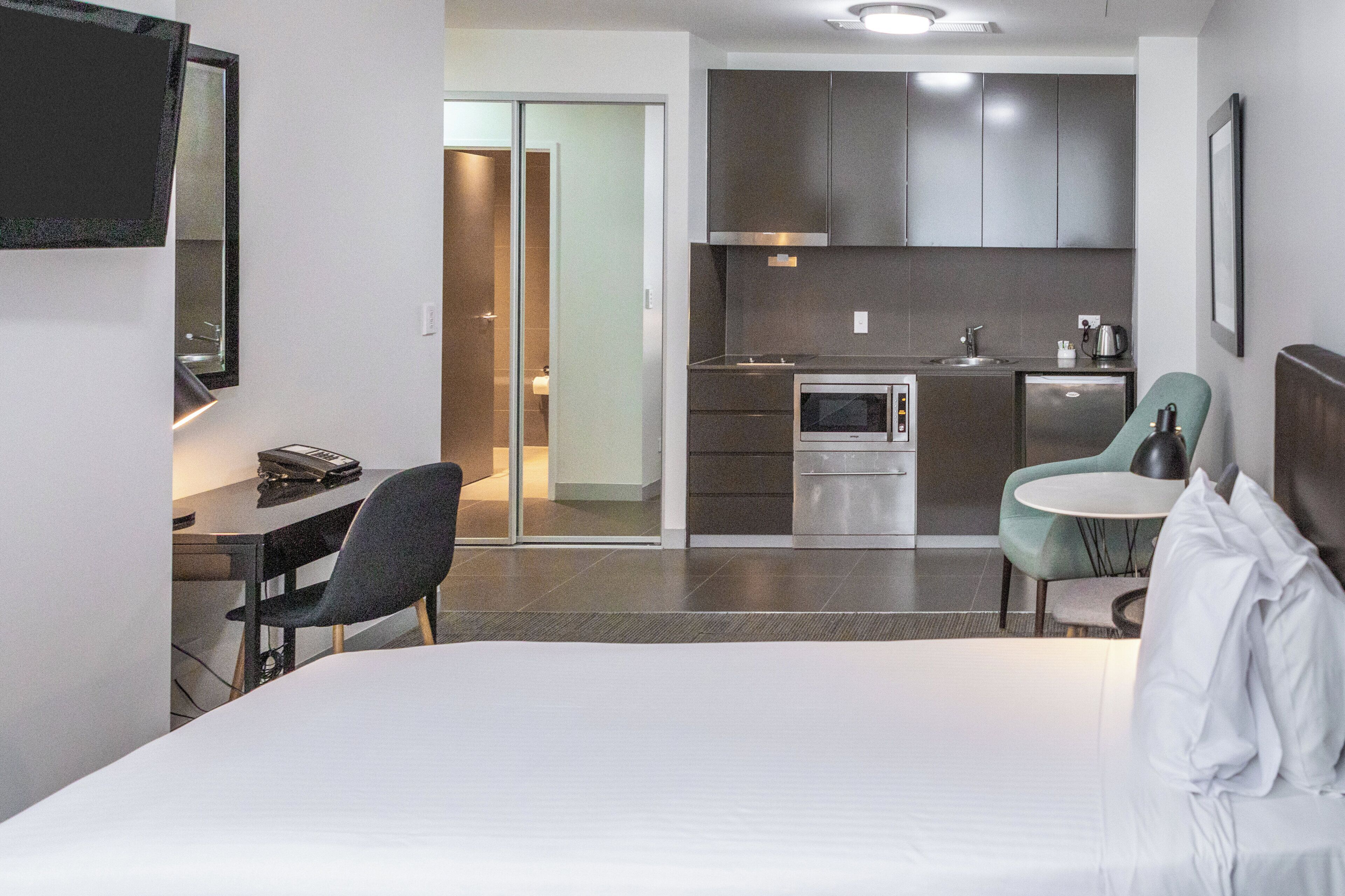 A hotel room with a bed, a desk, a chair, a kitchenette, and a dining area.