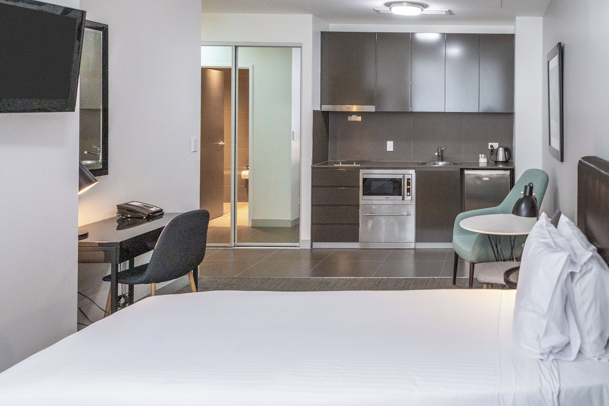 A hotel room with a bed, a desk, a chair, a kitchenette, and a dining area.