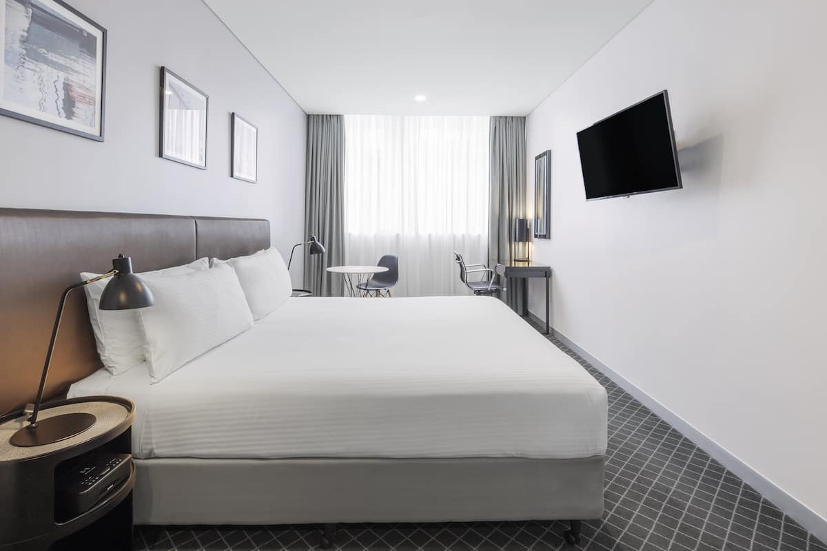 A modern hotel room with a large bed, bedside table, lamp, and a flat-screen TV mounted on the wall.