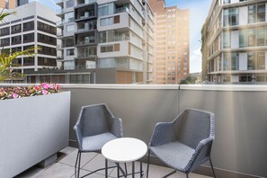 Un balcón con dos sillas de mimbre y una pequeña mesa redonda, con vista a un paisaje urbano de edificios modernos.