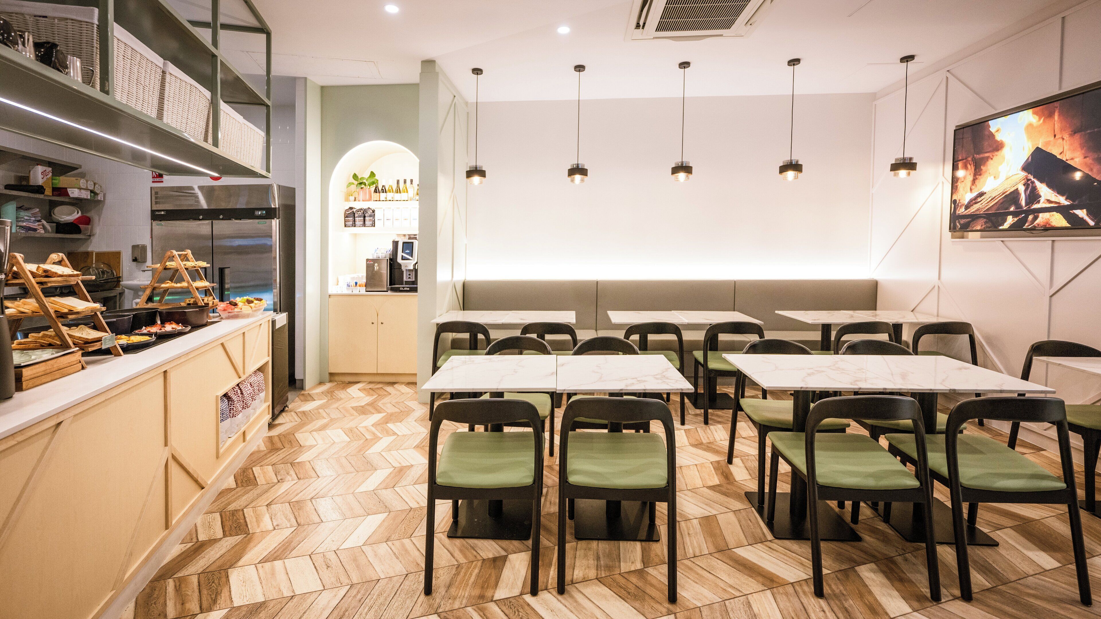 Een restaurantinterieur met een visgraat parketvloer, groene gestoffeerde stoelen, tafels met marmeren blad, een buffet met diverse gerechten, een koffiemachine en een flatscreen televisie waarop een vuurscène te zien is.