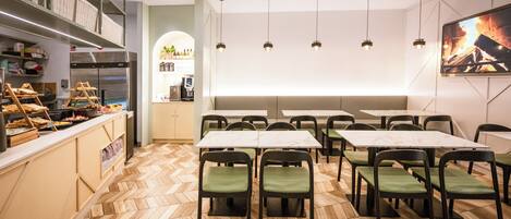 A restaurant interior with a herringbone wood floor, green cushioned chairs, marble-top tables, a buffet area with various food items, a coffee machine, and a flat-screen TV displaying a fire scene.