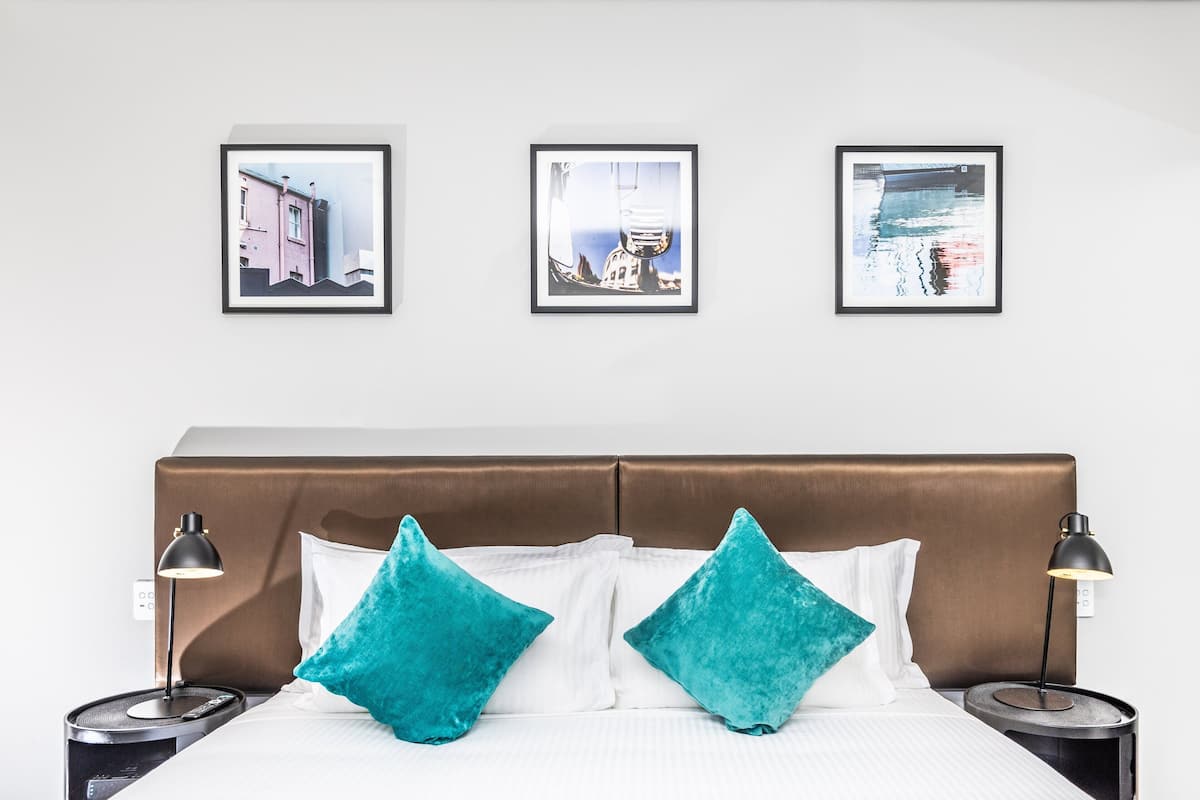 A hotel room with a brown headboard, white bedding, two teal pillows, and three framed pictures on the wall.