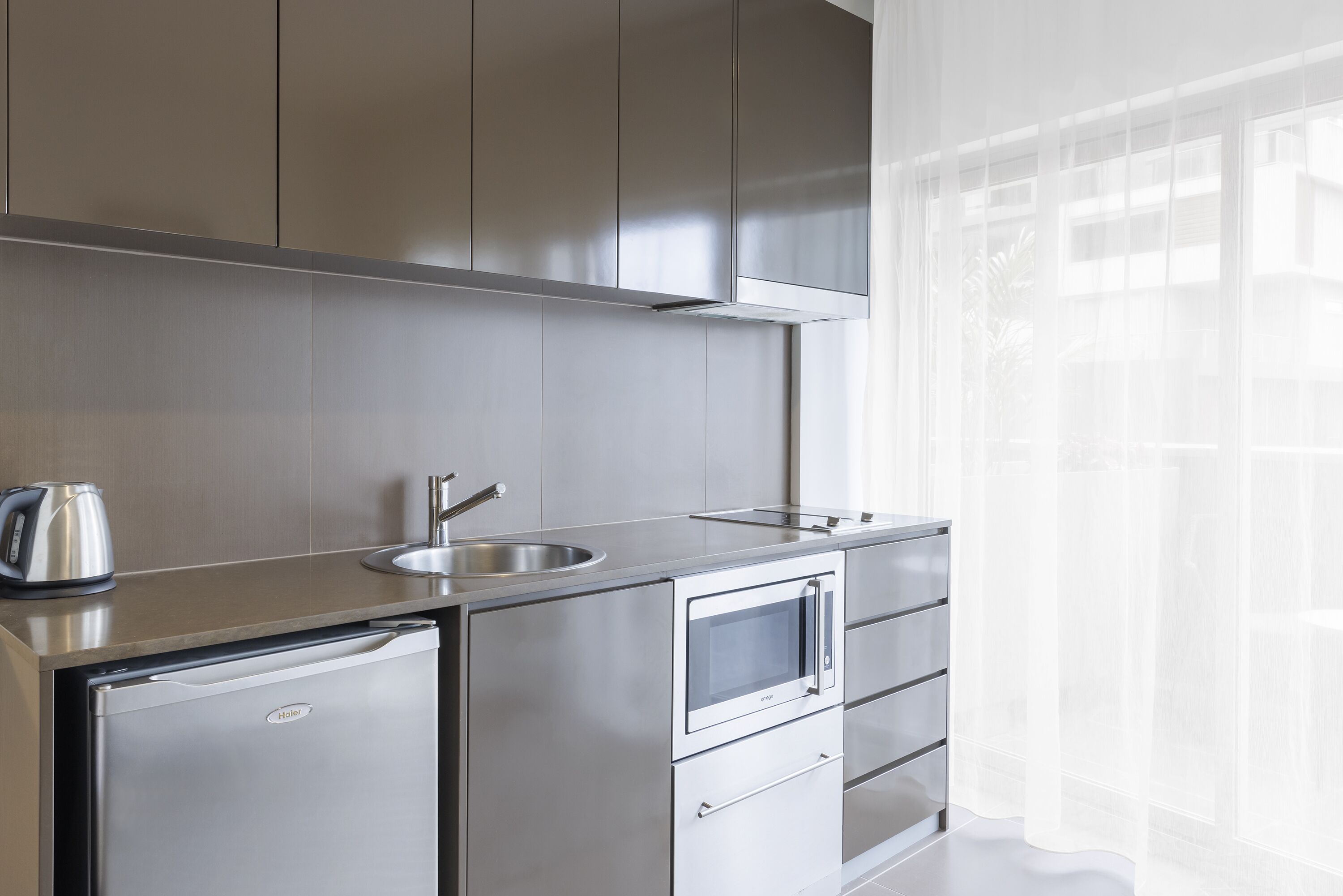 A modern kitchen with stainless steel appliances, a sink, and a window with sheer curtains.