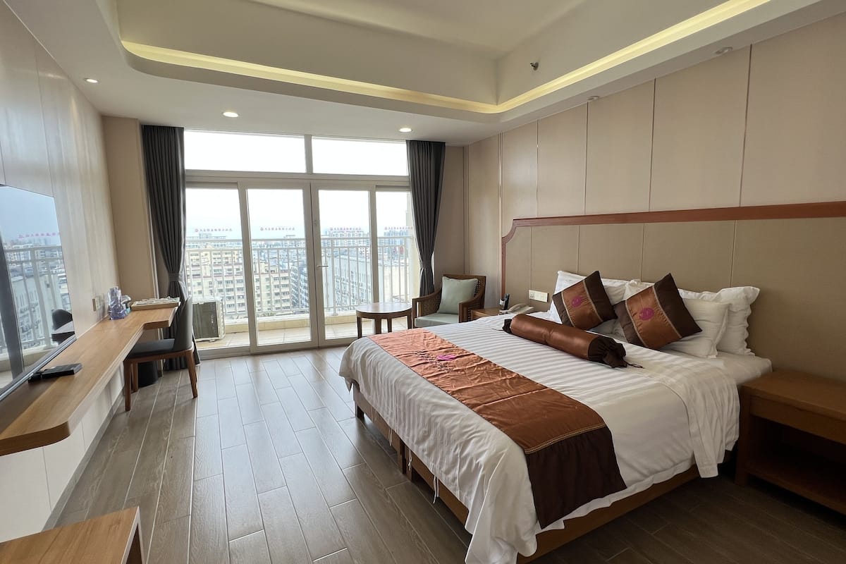 A hotel room with a large bed, a desk, a chair, a TV, and a view of the city through large windows.