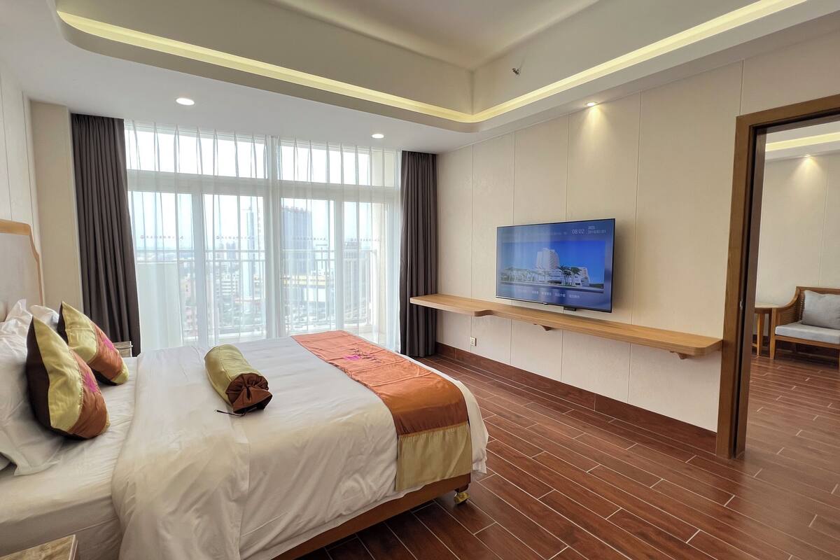 A hotel room with a large bed, a TV mounted on the wall, and a view of the city through large windows.