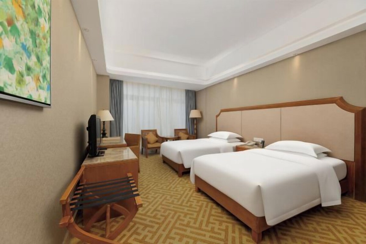 A hotel room with two beds, a desk with a chair, a TV, and a painting on the wall.