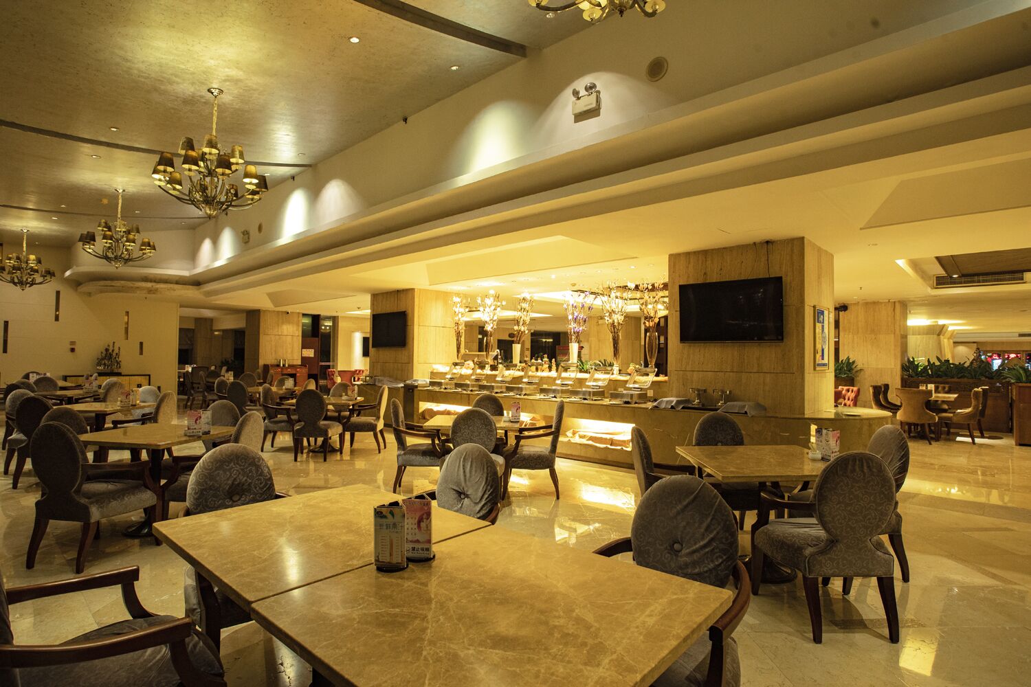 A spacious hotel lobby with multiple dining tables and chairs, a bar area, and a chandelier.