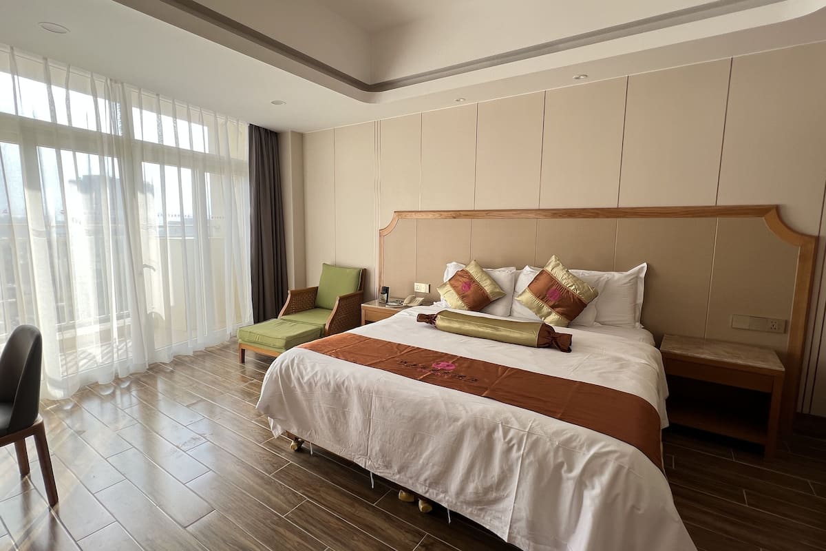 A hotel room with a large bed, a chair, a nightstand, and a window with sheer curtains.