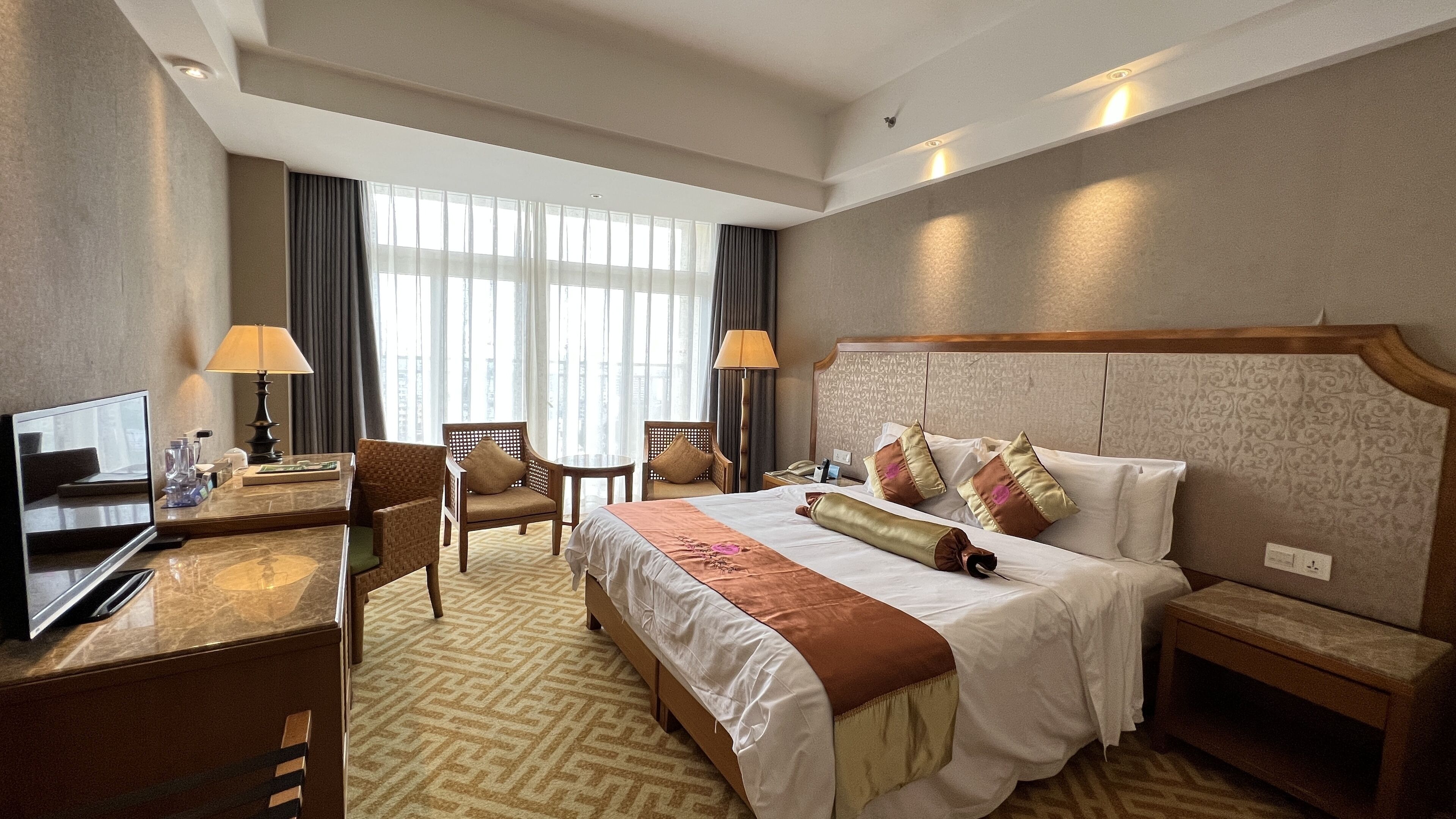 Quarto de hotel com uma cama grande, duas cadeiras, uma escrivaninha com computador e uma televisão.