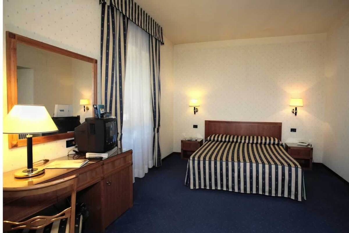 A hotel room with a bed, a desk with a lamp, a television, and two bedside tables with lamps.