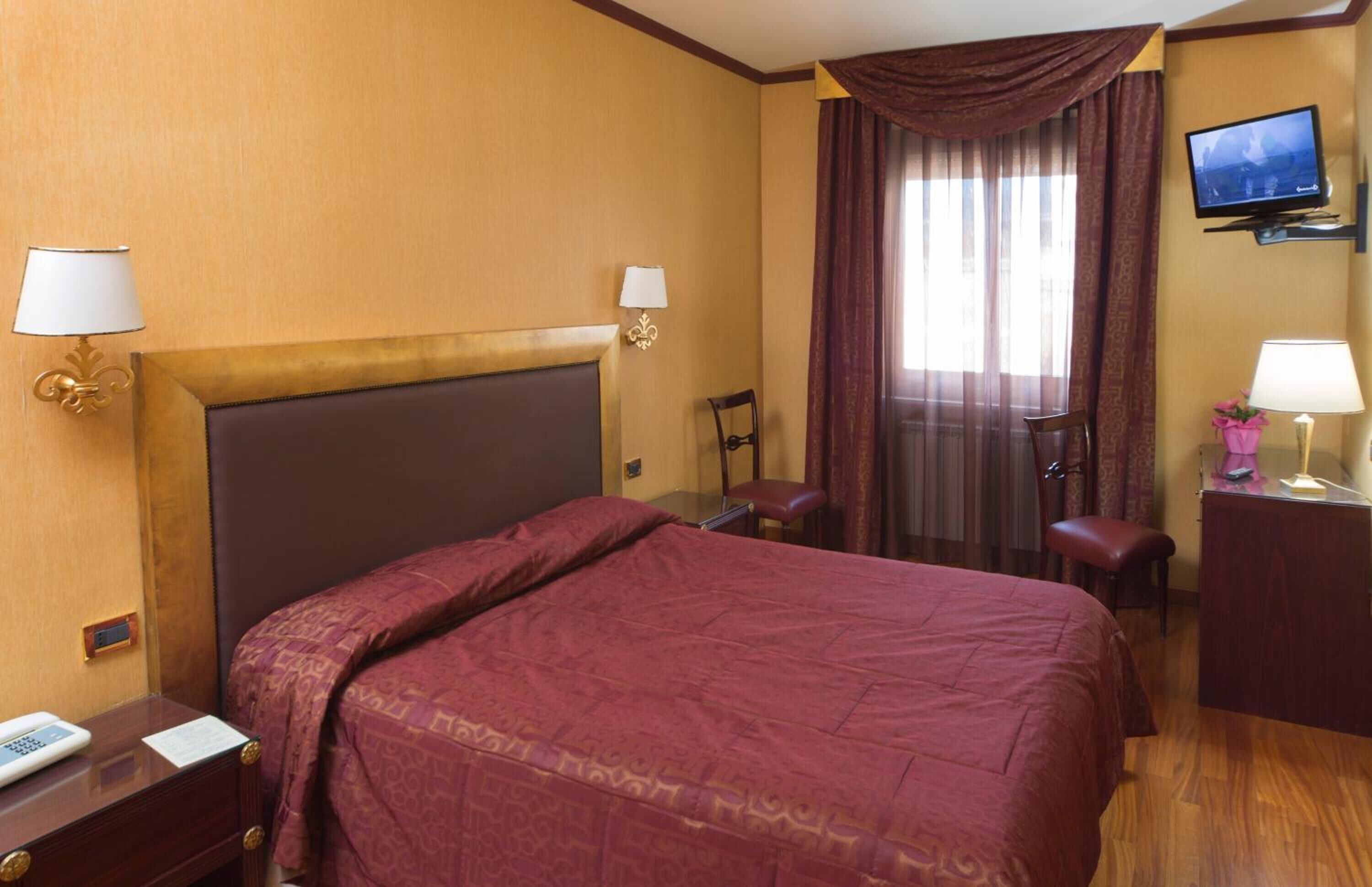Habitación de hotel con una cama grande, un escritorio con lámpara, una silla, televisor y una ventana con cortinas.