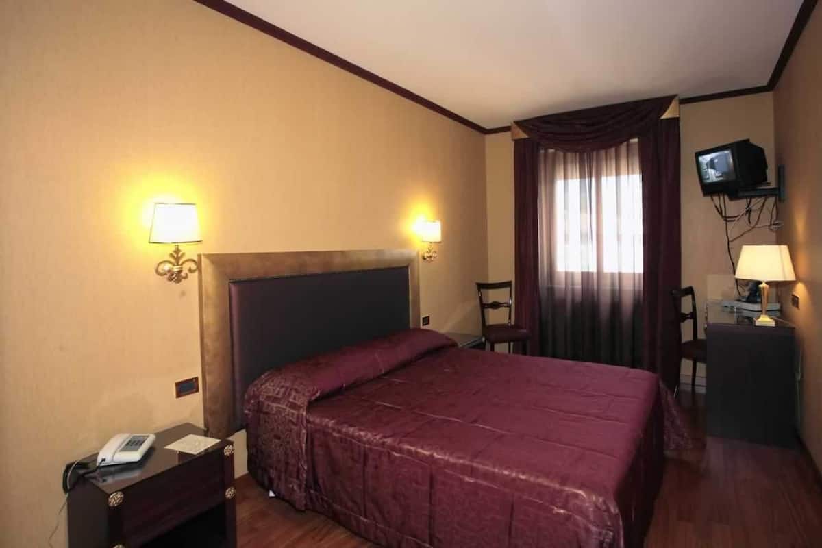 A hotel room with a large bed, a nightstand with a telephone, a television mounted on the wall, and a window with curtains.