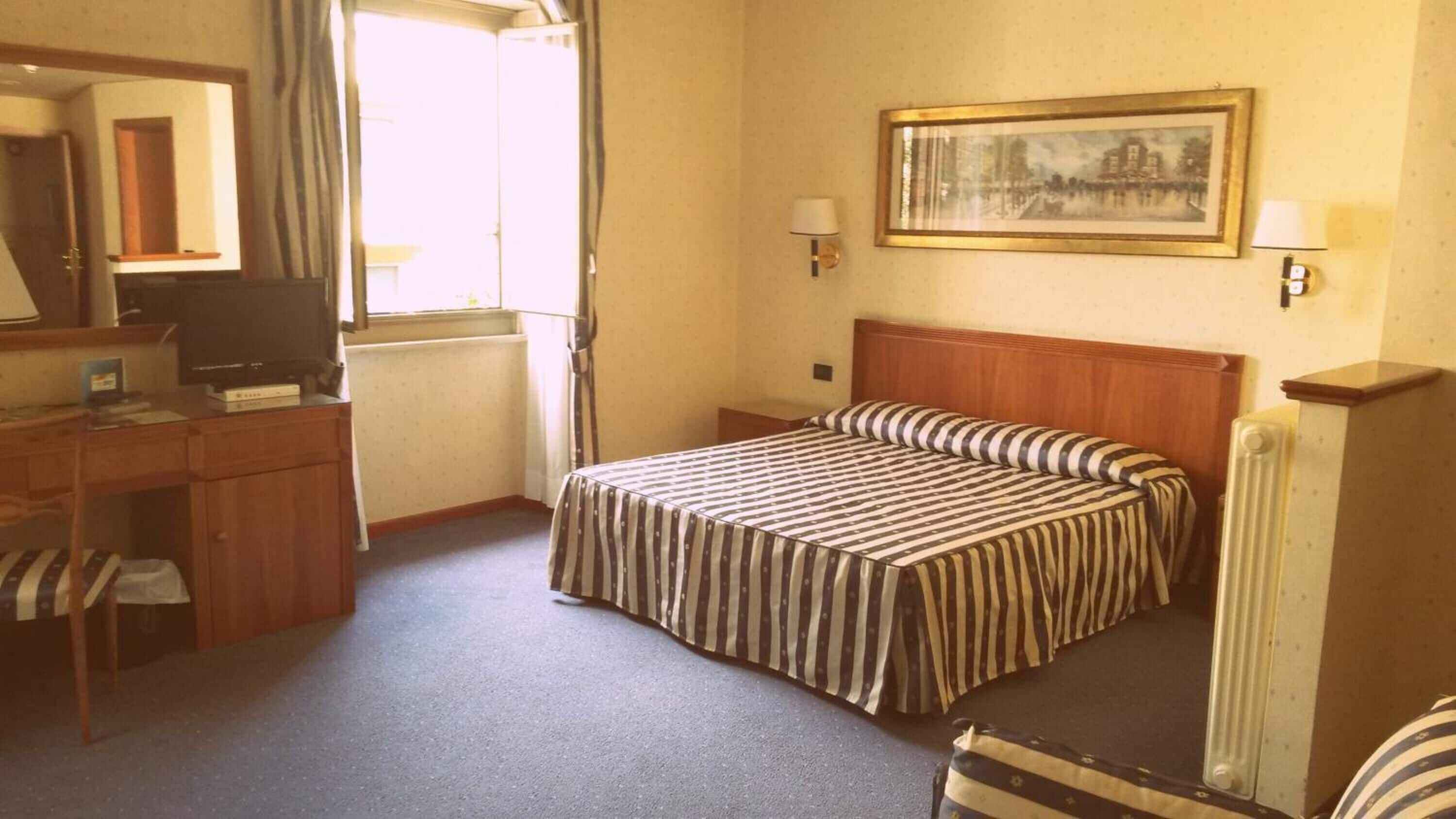 A hotel room with a bed, a desk, a chair, a television, a window, and a painting on the wall.