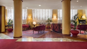 A spacious lobby with high ceilings, large columns, and a red carpet. There are several red upholstered chairs and a red sofa. The lobby has a patterned carpet and potted plants. - Hotel de Paris (Terni)