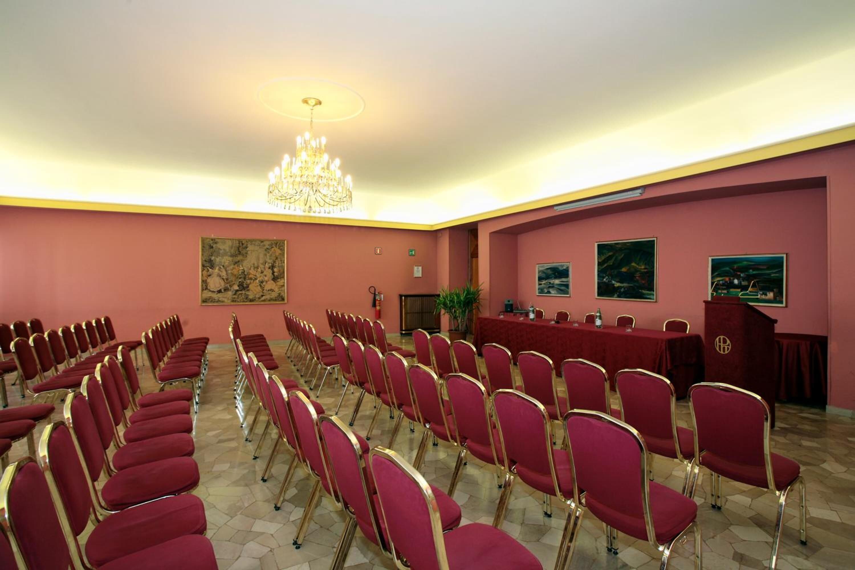 Uma sala de conferências com fileiras de cadeiras vermelhas, uma mesa comprida, um candelabro e quadros nas paredes.