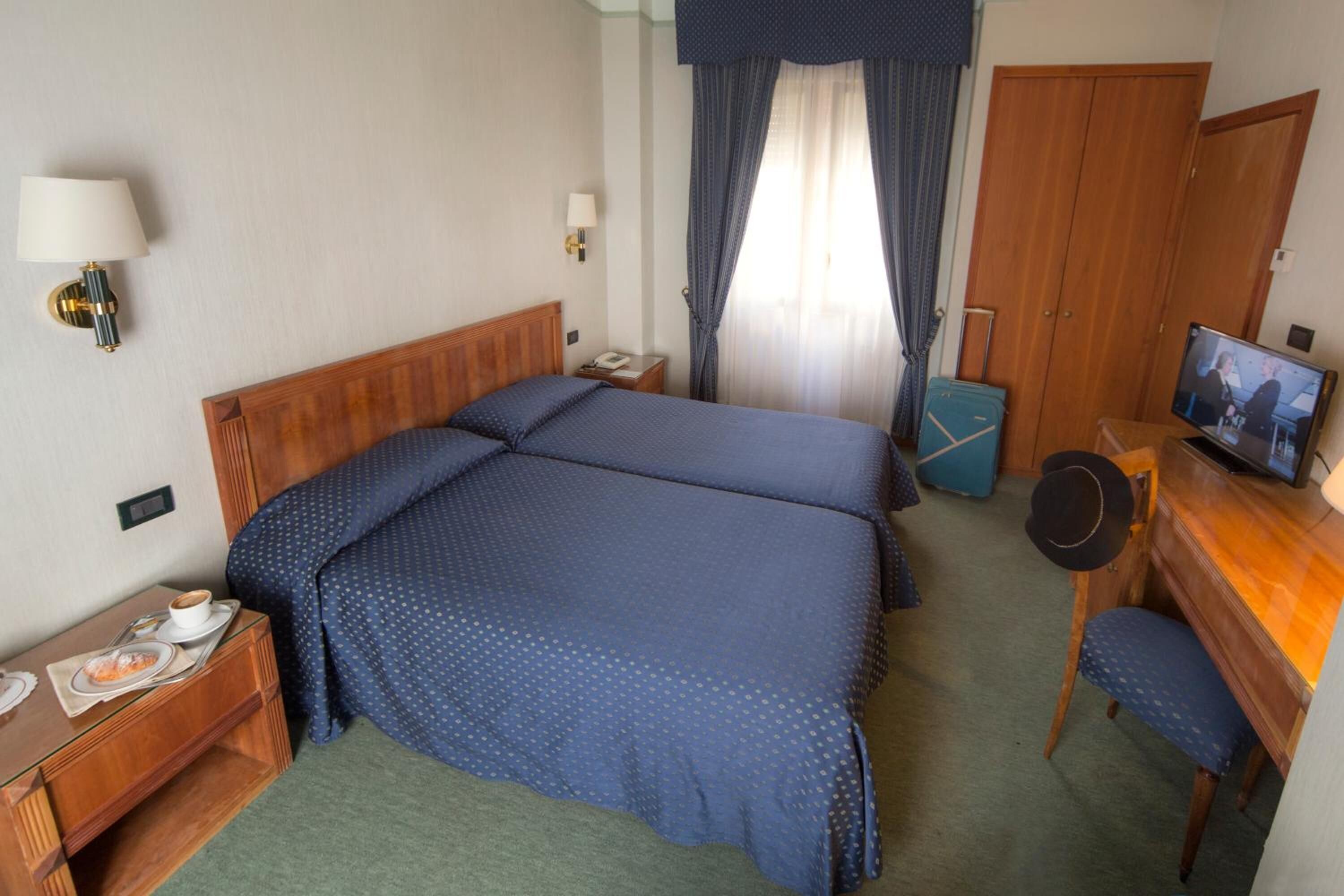 A hotel room with a large bed, a desk with a television, a chair, a desk lamp, a telephone, and a small table with a cup and a plate.