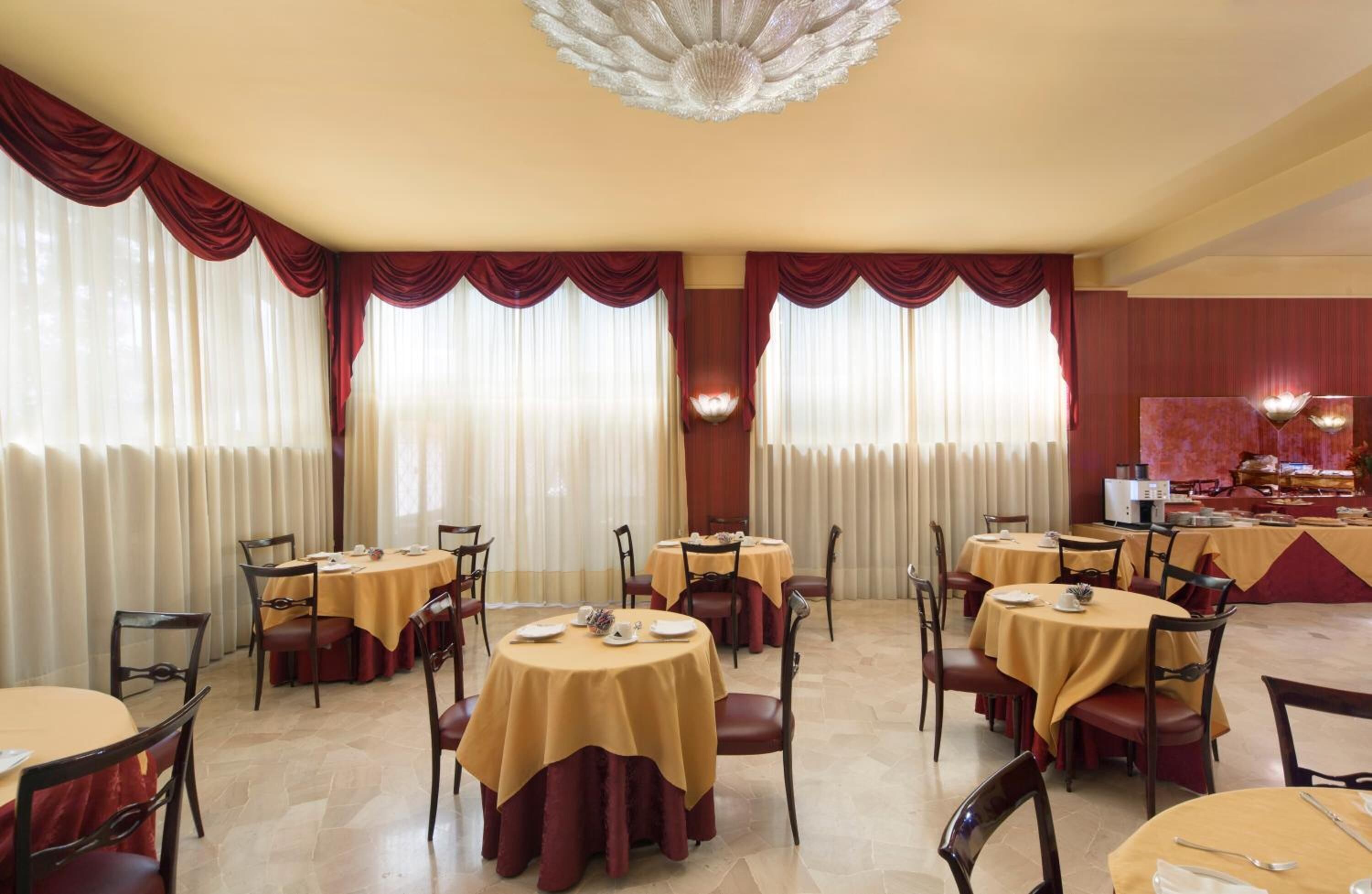 Un espace repas avec des tables rondes recouvertes de nappes jaunes, des rideaux rouges et un lustre.