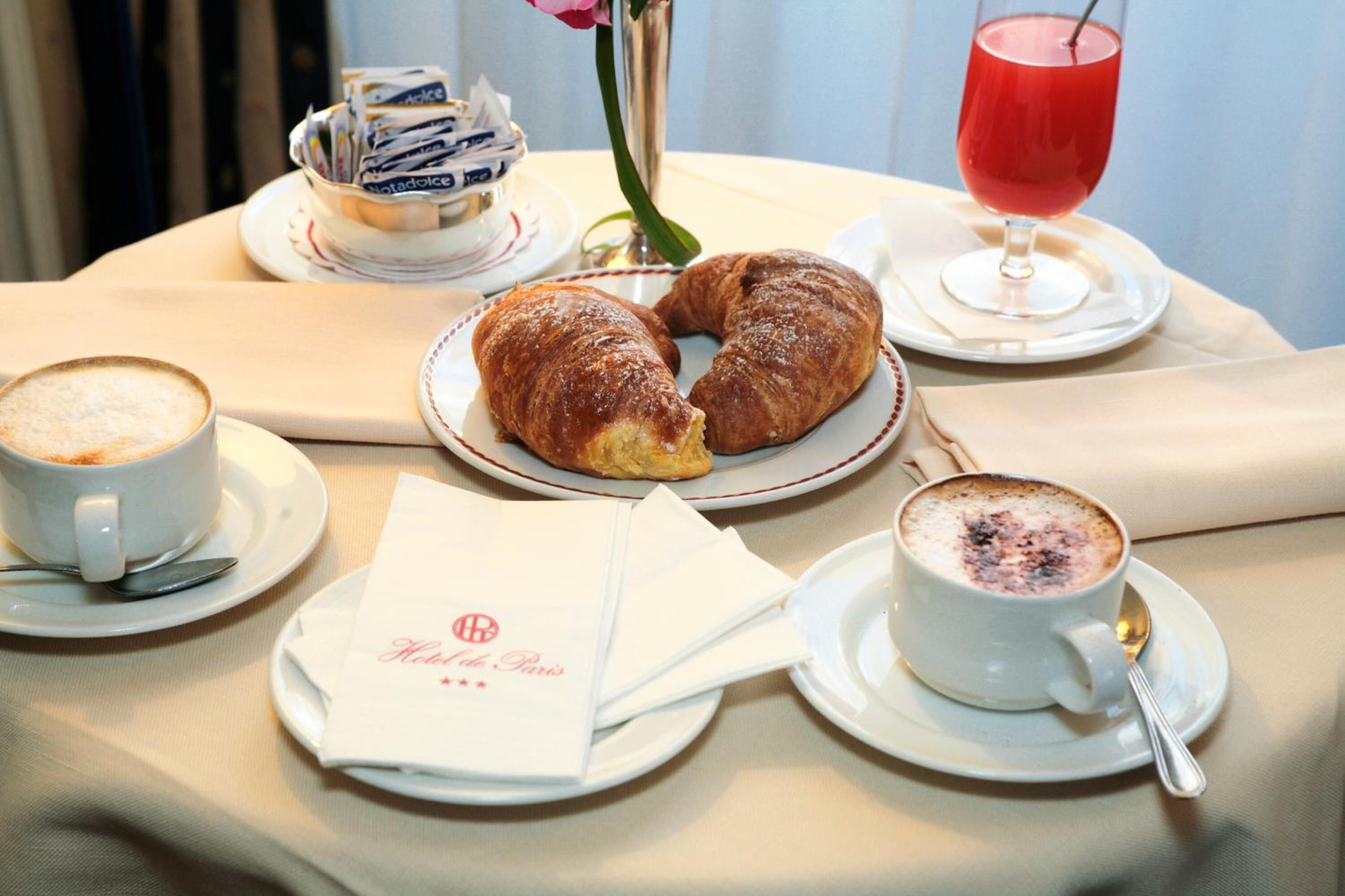 A table set with croissants, coffee, and a red drink, with a menu and a vase of flowers.