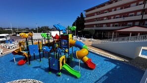 A children's play area with slides and climbing structures, a pool, and a building with balconies in the background.