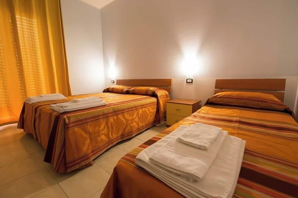Habitación de hotel con dos camas, cada una con cubrecama a rayas y cabecero de madera. Hay dos mesitas de noche, una junto a cada cama, y una mesita auxiliar pequeña entre las camas.