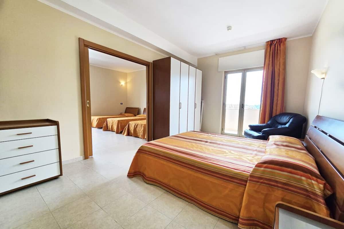 Habitación de hotel con una cama grande, un armario, un escritorio y una silla. Hay una puerta que da a otra habitación.