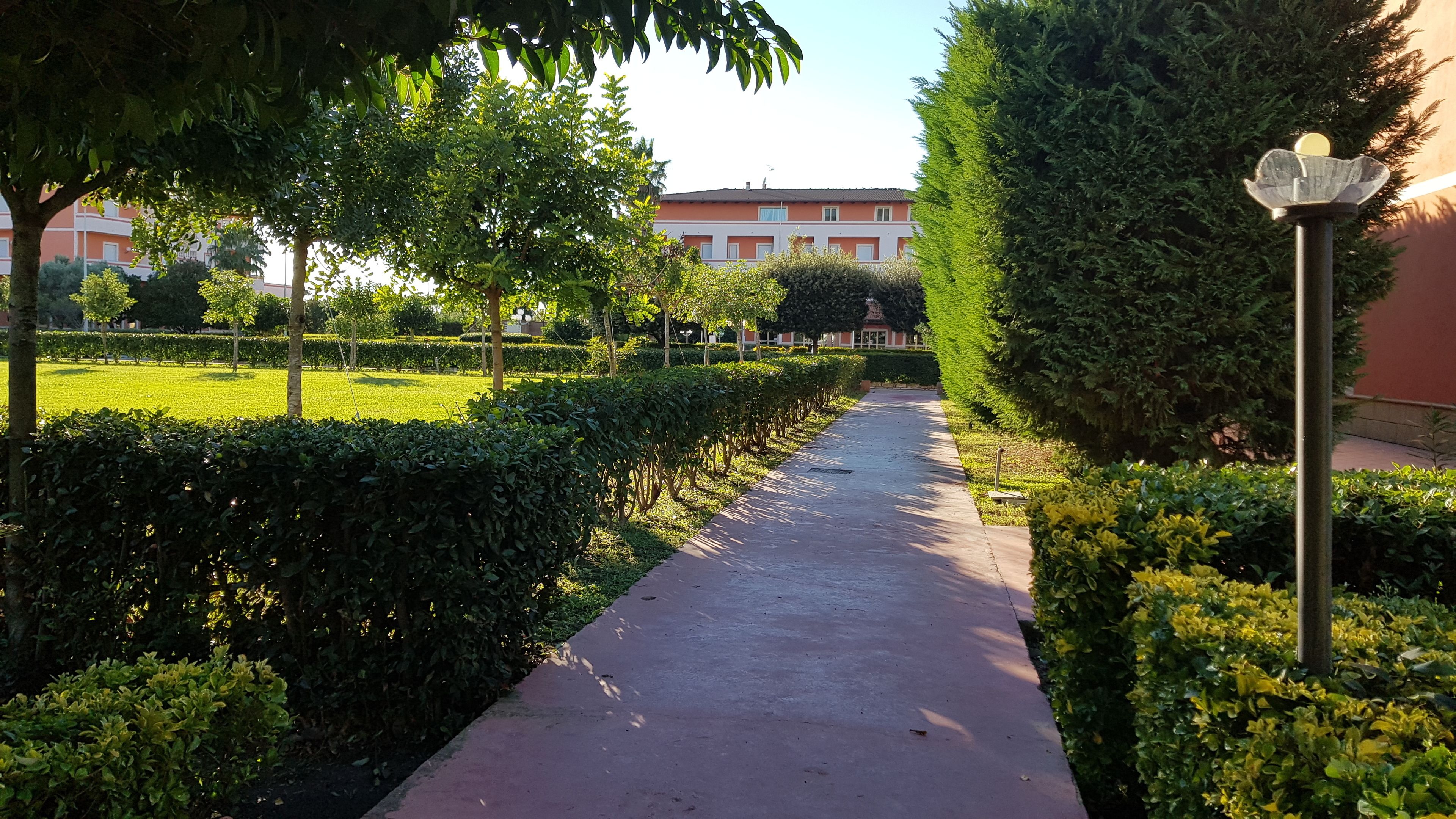 Um caminho ladeado por sebes e árvores bem aparadas, conduzindo a um edifício com fachada vermelha.