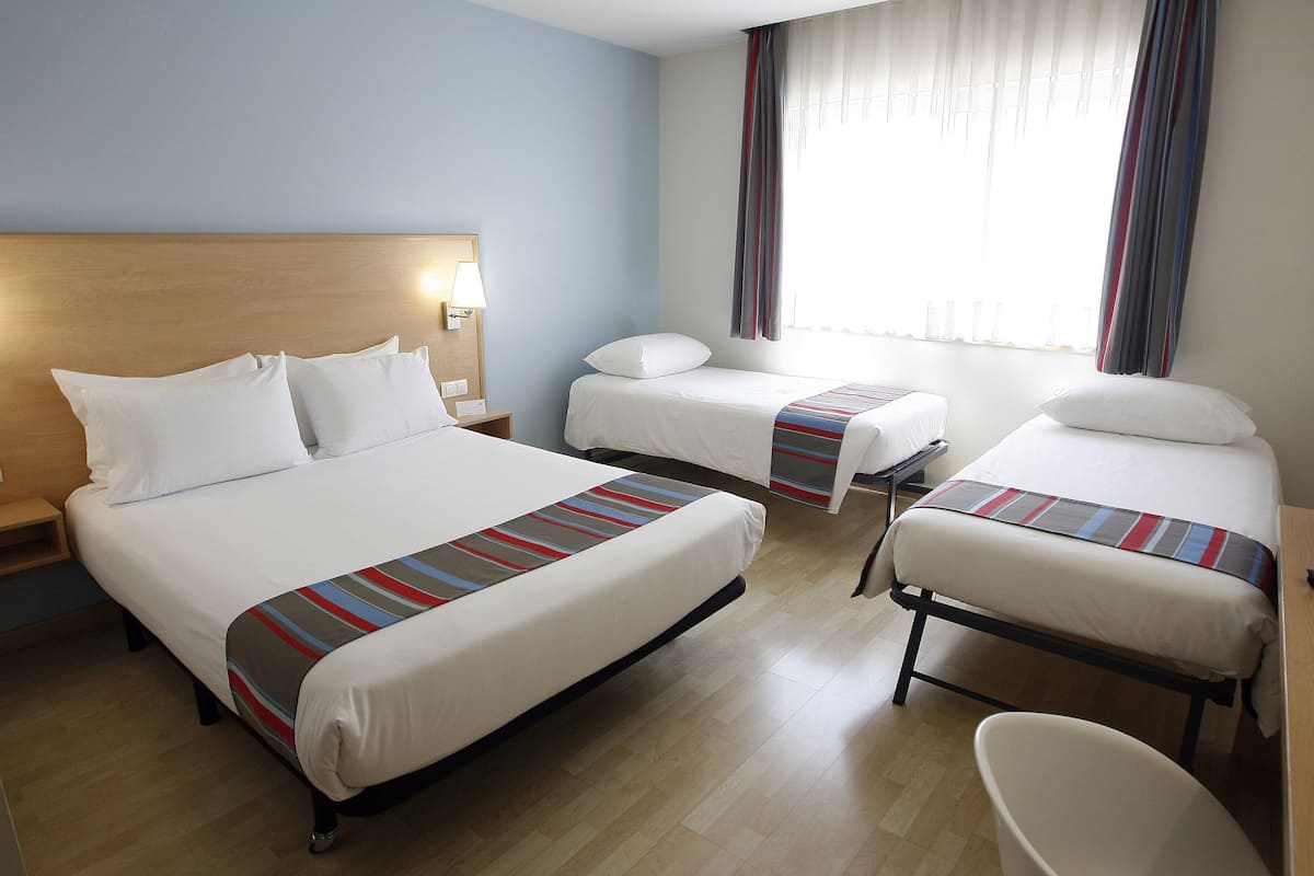 A hotel room with three beds, a wooden headboard, a nightstand, and a window with curtains.