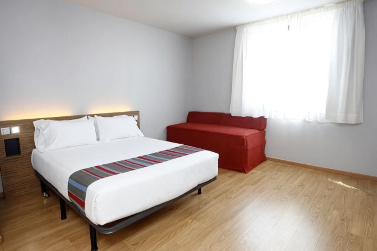 A hotel room with a bed, a red sofa, a nightstand, and a window with curtains.