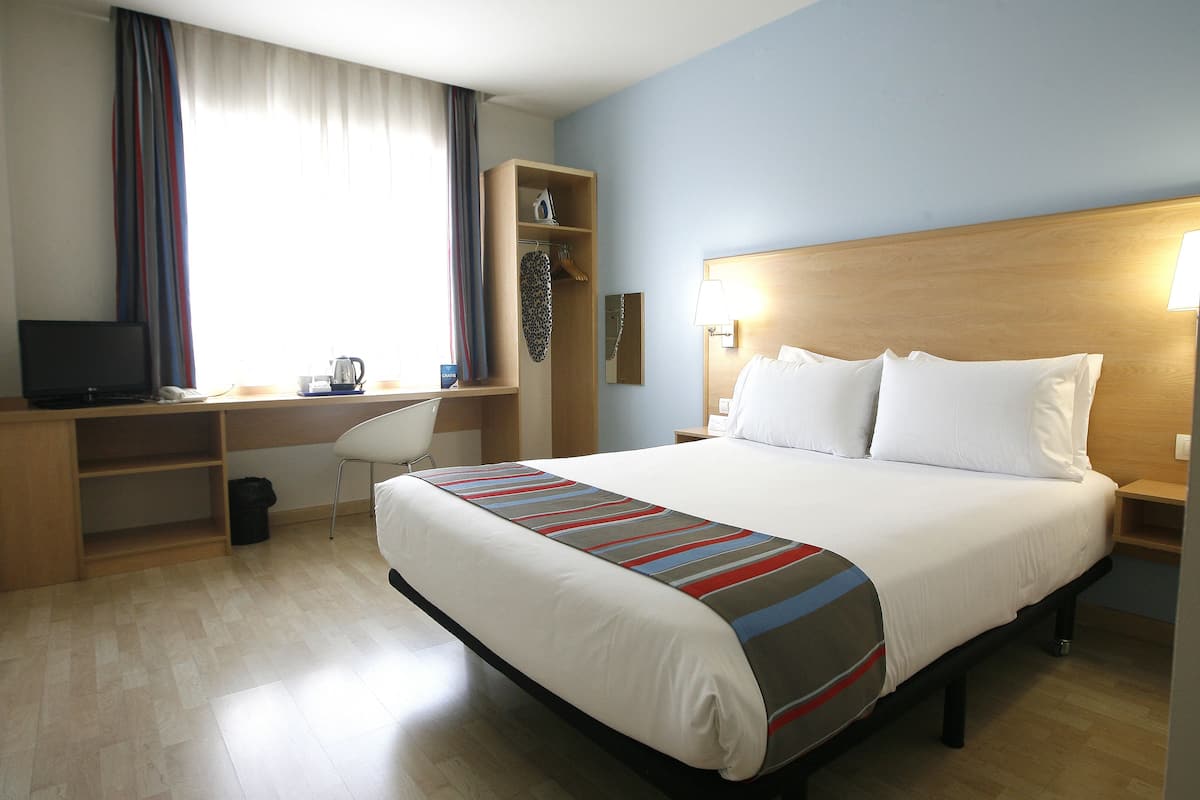 A hotel room with a bed, a desk with a chair, a television, and a window with curtains.