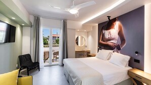 A modern hotel room with a large bed, a ceiling fan, a flat-screen TV, a desk, and a balcony with a view of buildings. - Thalero Holidays Center (Lefkada)